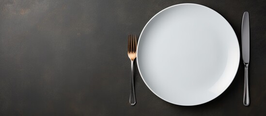 Copy space image of a white plate accompanied by a fork and spoon positioned on a sleek black slate surface in a flat lay arrangement