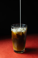 Milk pouring in glass with iced coffee on red and black background