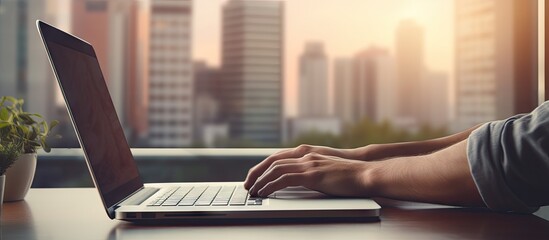 A close up of hands typing on a laptop placed on a wooden desk with a stack of papers nearby against the backdrop of a window The laptop screen is blank providing ample copy space