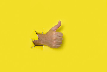 Hand with thumb up as a sign of approval, coming out of the hole of a yellow torn paper background.