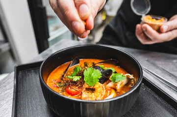 A chef’s hand garnishing a bowl of seafood soup with herbs, capturing the motion and precision in...