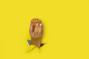 Female hand holding a cupcake coming out of a hole in a torn yellow paper background.