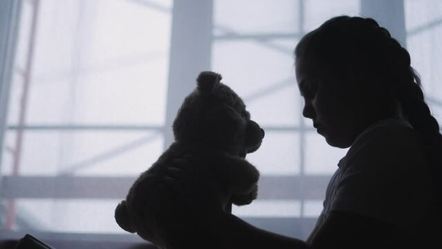 depressed girl child silhouette the window with her favorite toy teddy bear upset. problem of domestic violence in the family concept lifestyle. depressed girl is sad and bored at window with bear