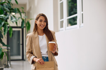 Young happy businesswoman with takeaway coffee and lunch bag at casual office looking at camera.