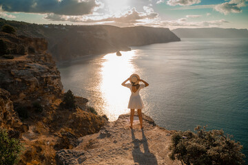 A woman stands on a cliff overlooking the ocean. She is wearing a white dress and a straw hat. The sky is cloudy and the sun is setting. - Powered by Adobe