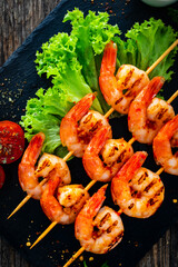 Grilled skewer shrimps and fresh vegetables served on black stony plate on wooden table
