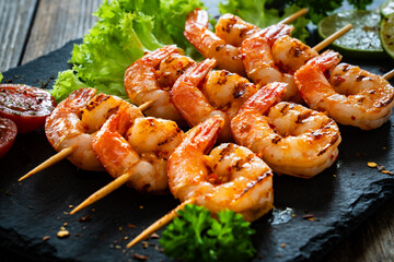 Grilled skewer shrimps and fresh vegetables served on black stony plate on wooden table