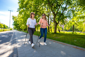 Nordic walking - two mid-adult beautiful women exercising in city park using Nordic walking poles 
