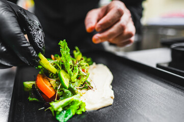 A close-up of hands wearing black gloves carefully plating a gourmet salad with mixed greens,...