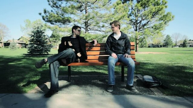 Footage of two young men sitting on a bench in park and talking to each other