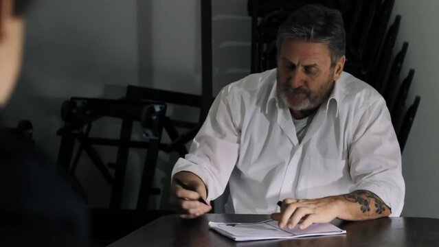 Old man with tattoos sitting and looking to another person behind the cam, then put a sign