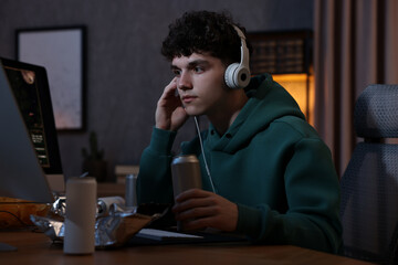 Young man with energy drink and headphones playing video game at wooden desk indoors