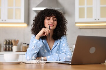 Beautiful young woman in stylish pyjama with pen using laptop at wooden table in kitchen
