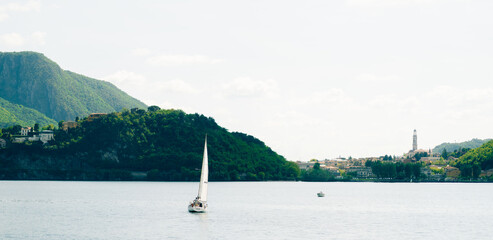 sailboat on lake with green mountains and village in the distance under cloudy sky