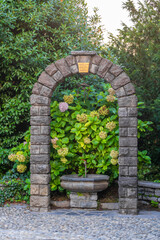 rustic stone arch framing a small fountain surrounded by blooming hydrangeas in a lush garden