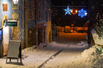 snow-covered street at night with festive lights and restaurant sign, creating a cozy winter scene