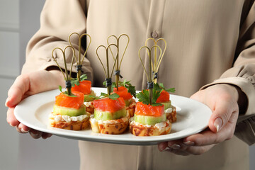 Woman holding tasty canapes with salmon, cucumber, bread and cream cheese on grey background,...