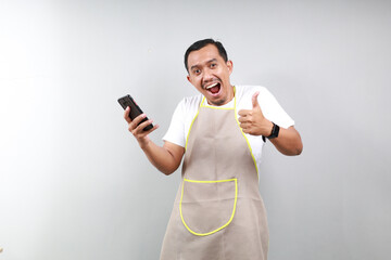 Satisfied happy Asian man barista barman employee wear brown apron hold in hand use mobile cell...