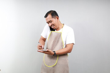 Handsome asian barista in apron is talking on the mobile phone, making notes and smiling while...