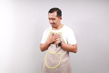 Relieved Handsome asian barista in apron standing while holding chest. Isolated on gray background