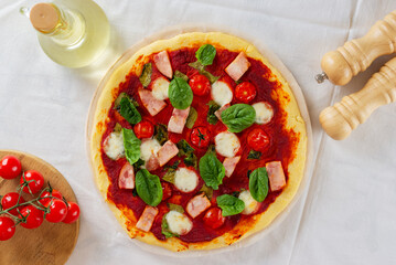 Handmade, DIY pizza with ham, tomatoes, mozzarella, green, sauce, spices and oil on a table in kitchen. Homemade pizza. Top view.