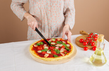 Woman cutting fresh pizza, handmade, DIY pizza with ham, tomatoes, mozzarella and green on a table. Homemade pizza, yeast-free dough.
