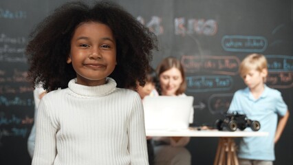 African girl smiling and looking at camera while multicultural friends working or learning engineering code or prompt in STEM technology classroom from young beautiful caucasian teacher. Erudition. - Powered by Adobe