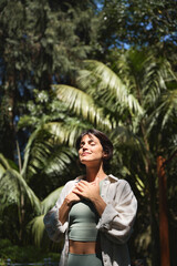 Happy calm young Hispanic woman holding hands on chest meditating doing yoga breathing exercises with eyes closed feeling gratitude, mental balance standing in green nature tropical park. Vertical.