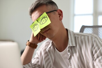 Man with fake eyes painted on sticky notes snoozing at workplace in office