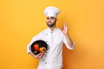 Professional chef holding colander with vegetables and showing OK gesture on yellow background