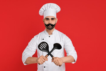Naklejka premium Professional chef with funny artificial moustache holding kitchen utensils on red background