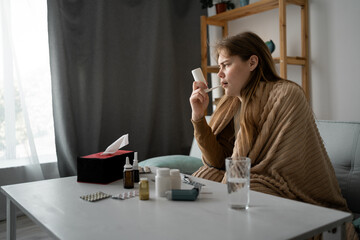 side view of a young Caucasian girl sick sitting at home on the sofa, wrapped in a blanket, using a throat spray for cough. The concept of treating viruses for a healthy lifestyle in the cold season
