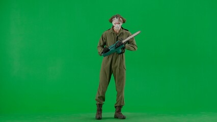 A maniac in a hockey mask and overalls with a chainsaw in his hands. Unrecognizable man in scary Jason Voorhees look on green screen chromakey. Halloween, horror.