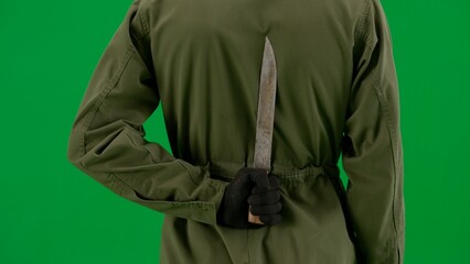 Fototapeta premium Rear view of a maniac hiding a knife behind his back. Man in jumpsuit with weapon on green screen chroma key close up. Halloween, horror.