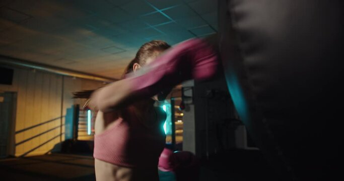Young woman throwing a powerful punch at a heavy bag in a dimly lit gym with an illuminated boxing ring, showcasing intense training and fitness dedication