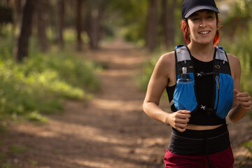 A woman is running through a forest with a blue backpack on. She is smiling and she is enjoying her...