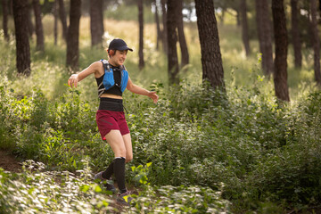 A woman is running through a forest with a backpack on. She is wearing a red shirt and shorts