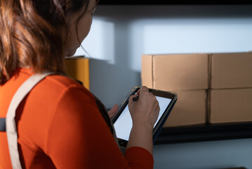 A woman uses a digital tablet to check stock inventory at home, She stands next to shelves with...