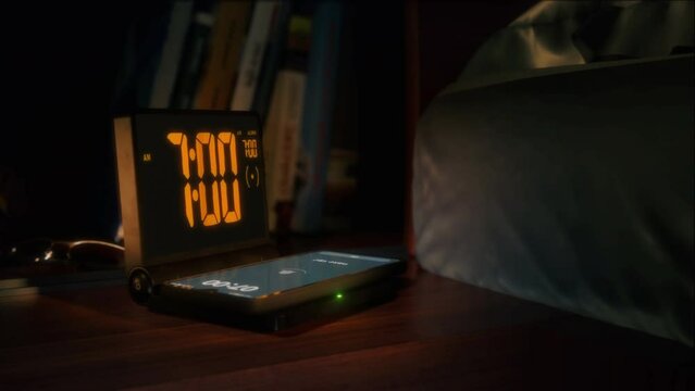 Digital alarm clock with phone charger and orange clockface is waking up at 7 AM. The numbers on the clock screen changes from 6:59 no 7:00 AM. And the alarm goes off on the phone that is charging.