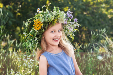 Happy Children Holiday on Nature.  Smiling Child with Flower Wreath in Summer Holidays. Blonde Little Girl with Flower Wreath on Green Nature Background. Solstice Midsummer Concept. 