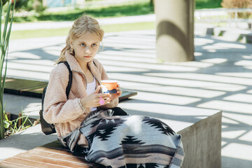 A little girl eats sweets from a cup, sitting on a bench.