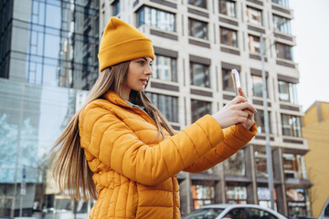A beautiful woman in a mustard hat and jacket takes a selfie on the phone against the background of a modern city building.