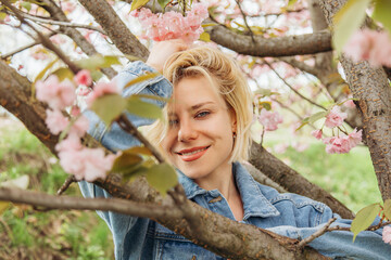 Close-up portrait of a young blonde woman among branches with blooming cherry blossoms.