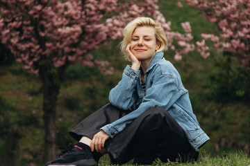 A smiling young blonde is sitting on the grass against the background of blossoming sakura trees.