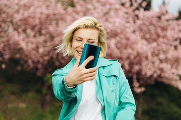A young happy woman in a turquoise trench coat taking a selfie while enjoying the cherry blossoms in the park.