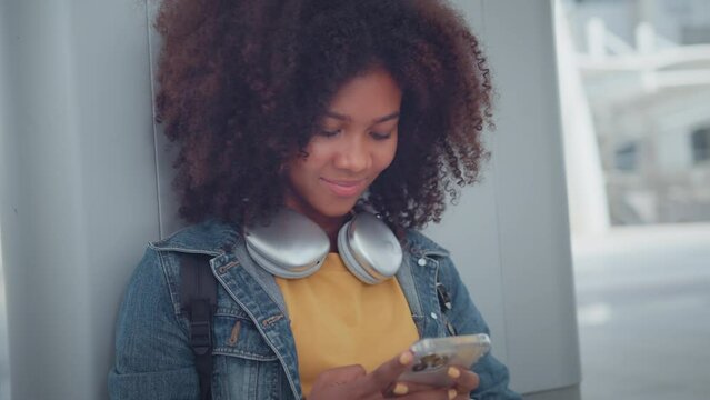 Smiling african american woman wearing headphones listening to music online from smartphone in the urban city. Female listening to music, song, podcast, or audiobook.