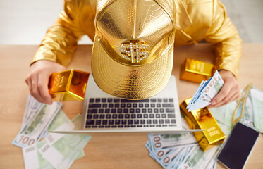 Top view of young funny man sitting at desk, holding gold bar and dollars banknotes, working online...