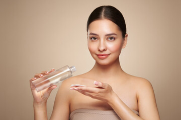 Smiling woman holds bottle with cosmetic tonic. Photo of attractive woman with perfect makeup on beige background. Beauty and skin care concept. Removing makeup