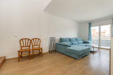 Living room with a large sofa upholstered in blue fabric, some varnished wooden chairs and a balcony with matching curtains