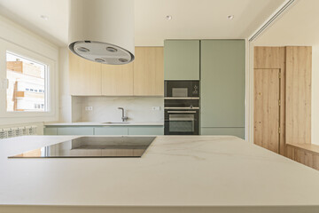 an open kitchen with a modern design with pastel green furniture, with a matching island with white marble countertops and a window on the left side